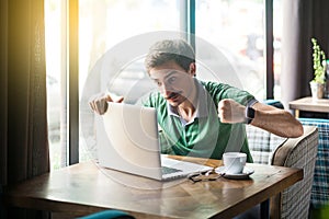 Young angry businessman in green t-shirt sitting with aggressive face and boxing fists and try to punching his laptop