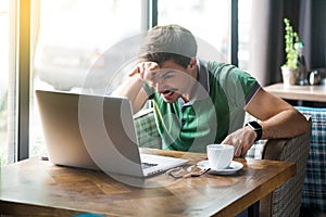 Young angry businessman in green t-shirt sitting and looking at laptop display with crazy face and horns gesture on his forehead
