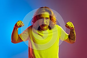 Young angry man with long curly hair posing  on gradient blue-pink background. Concept of beauty, fashion, youth