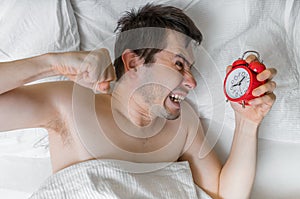 Young angry man is waking up and wants to damage his alarm clock with fist