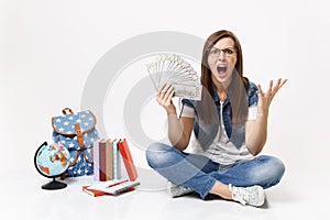 Young angry woman student screaming spreading hands holding bundle lots of dollars, cash money sit near globe backpack