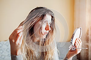 Young annoyed woman is upset about dirty oily and greasy hair looking in mirror at home. Health treatment