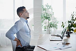 Young Arab businessman having acute back pain, massaging aching muscles, sitting at desk in modern office, copy space