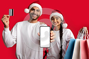 Young Arab Couple In Santa Hats Holding Blank Smartphone And Credit Card