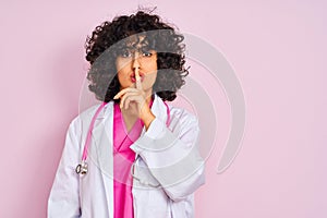 Young arab doctor woman with curly hair wearing stethoscope over isolated pink background asking to be quiet with finger on lips