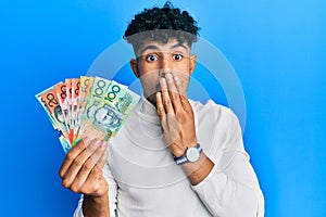 Young arab handsome man holding australian dollars covering mouth with hand, shocked and afraid for mistake