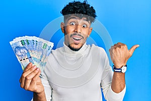 Young arab handsome man holding south african 100 rand banknotes pointing thumb up to the side smiling happy with open mouth