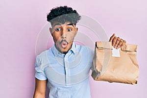 Young arab handsome man holding take away paper bag scared and amazed with open mouth for surprise, disbelief face