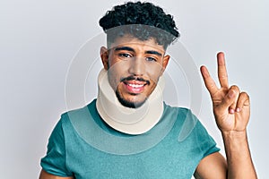 Young arab handsome man wearing cervical neck collar smiling with happy face winking at the camera doing victory sign