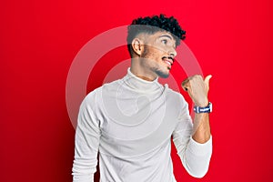 Young arab handsome man wearing turtleneck sweater pointing thumb up to the side smiling happy with open mouth
