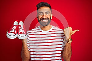 Young arab indian hispanic man holding sport casual sneakers over isolated red background pointing and showing with thumb up to