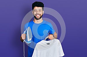 Young arab man with beard holding electric steam iron and white t shirt smiling and laughing hard out loud because funny crazy