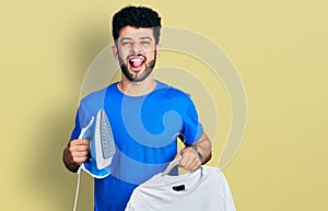 Young arab man with beard holding electric steam iron and white t shirt sticking tongue out happy with funny expression