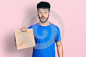 Young arab man with beard holding take away paper bag scared and amazed with open mouth for surprise, disbelief face