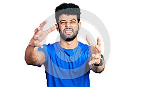 Young arab man with beard wearing casual blue t shirt shouting frustrated with rage, hands trying to strangle, yelling mad