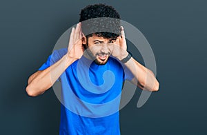 Young arab man with beard wearing casual blue t shirt trying to hear both hands on ear gesture, curious for gossip