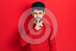 Young arab man with beard wearing casual red sweater asking to be quiet with finger on lips