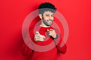 Young arab man with beard wearing casual red sweater pointing fingers to camera with happy and funny face