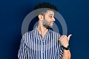Young arab man with beard wearing casual striped shirt pointing thumb up to the side smiling happy with open mouth