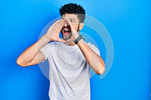 Young arab man with beard wearing casual white t shirt shouting angry out loud with hands over mouth