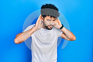 Young arab man with beard wearing casual white t shirt trying to hear both hands on ear gesture, curious for gossip