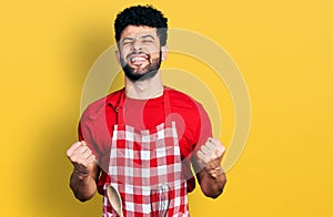 Young arab man with beard wearing cook apron excited for success with arms raised and eyes closed celebrating victory smiling