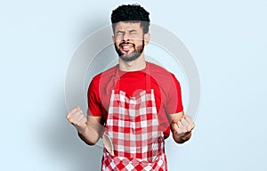 Young arab man with beard wearing cook apron very happy and excited doing winner gesture with arms raised, smiling and screaming