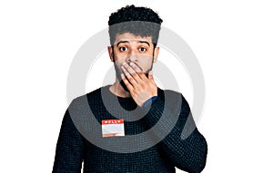 Young arab man with beard wearing hello my name is sticker identification covering mouth with hand, shocked and afraid for mistake