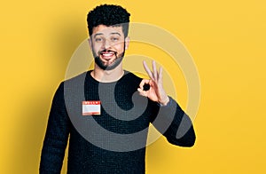 Young arab man with beard wearing hello my name is sticker identification doing ok sign with fingers, smiling friendly gesturing