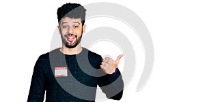 Young arab man with beard wearing hello my name is sticker identification pointing thumb up to the side smiling happy with open