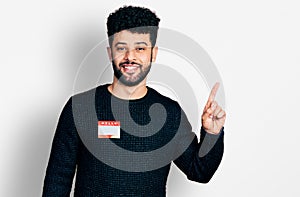 Young arab man with beard wearing hello my name is sticker identification smiling happy pointing with hand and finger to the side