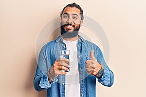 Young arab man drinking glass of water smiling happy and positive, thumb up doing excellent and approval sign