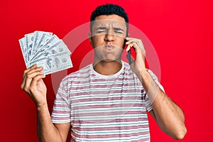 Young arab man having conversation talking on the smartphone holding dollars puffing cheeks with funny face