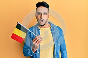 Young arab man holding belgium flag scared and amazed with open mouth for surprise, disbelief face