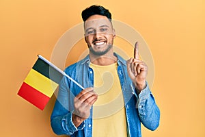 Young arab man holding belgium flag smiling with an idea or question pointing finger with happy face, number one