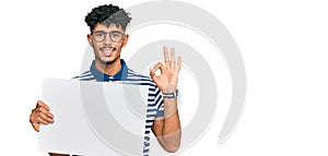 Young arab man holding blank empty banner doing ok sign with fingers, smiling friendly gesturing excellent symbol