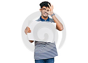 Young arab man holding blank empty banner smiling happy doing ok sign with hand on eye looking through fingers