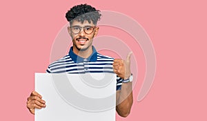 Young arab man holding blank empty banner smiling happy and positive, thumb up doing excellent and approval sign