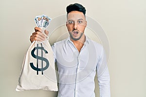 Young arab man holding dollars bag scared and amazed with open mouth for surprise, disbelief face