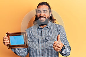 Young arab man holding empty frame smiling happy and positive, thumb up doing excellent and approval sign