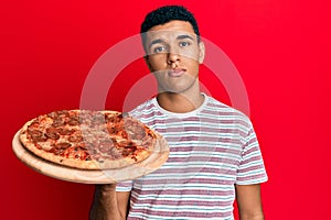 Young arab man holding italian pizza thinking attitude and sober expression looking self confident