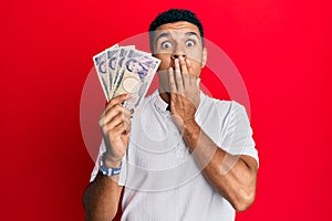 Young arab man holding japanese yen banknotes covering mouth with hand, shocked and afraid for mistake