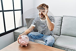 Young arab man holding piggy bank smiling happy doing ok sign with hand on eye looking through fingers