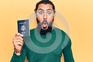 Young arab man holding united states passport scared and amazed with open mouth for surprise, disbelief face