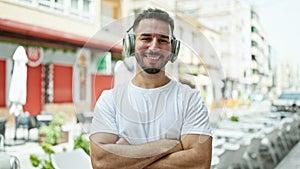 Young arab man listening to music standing with arms crossed gesture at coffee shop terrace