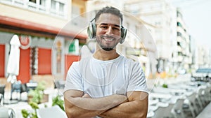 Young arab man listening to music standing with arms crossed gesture at coffee shop terrace