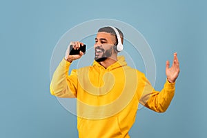 Young arab man singing, holding smartphone like microphone wearing wireless headphones and having fun, blue background