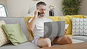 Young arab man talking on smartphone using laptop reading paper at home