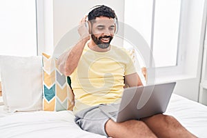 Young arab man using laptop and headphones sitting on bed at bedroom