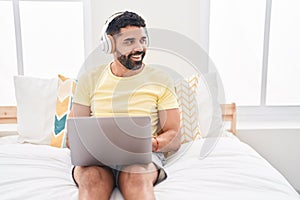 Young arab man using laptop and headphones sitting on bed at bedroom